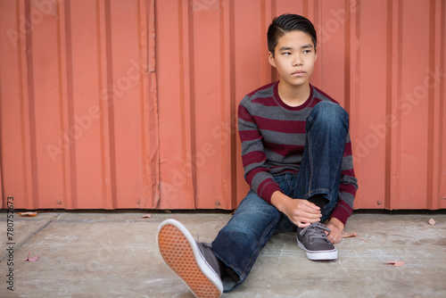Sad teen Asian boy sitting against a wall looking pensive and off the the left.