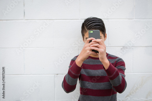 Teenage asian boy standing alone against an outdoor white background, holding his smartphone up to obscure his face