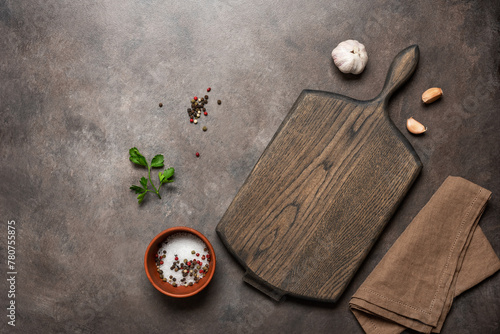 Culinary background with empty wooden cutting board, spices and garlic, dark brown rustic table. Top view, flat lay, copy space.