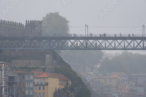 People with umbrellas and raincoats walking on the upper platform of the Don Luis I steel bridge with the medieval wall below, the funicular and the old town of the city of Porto in Portugal.