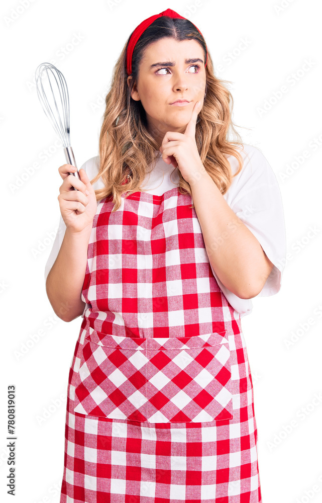 Young caucasian woman wearing apron holding whisk serious face thinking about question with hand on chin, thoughtful about confusing idea Young caucasian woman wearing apron holding whisk serious face thinking about question with hand on chin, thoughtful about confusing idea