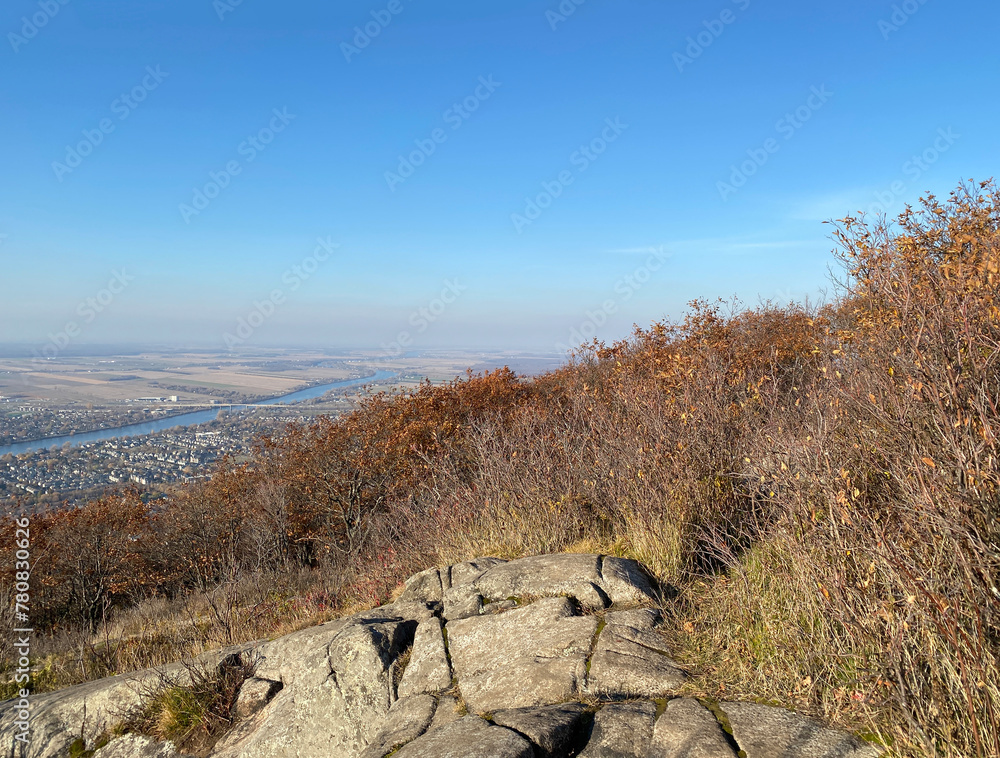 Autumn in the mountains. Mountain top with view of city, river, roads, houses, trees and blue sky. Landscape on a summit of a mountain. Rock and dry bushes in foreground of panoramic aerial view. Autumn in the mountains. Mountain top with view of city, river, roads, houses, trees and blue sky. Landscape on a summit of a mountain. Rock and dry bushes in foreground of panoramic aerial view.