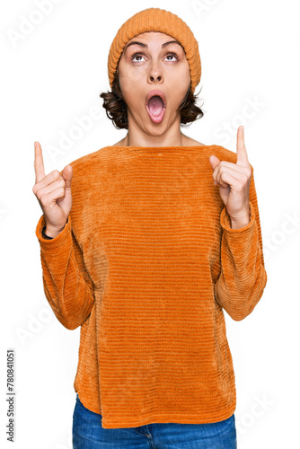Young hispanic woman wearing casual clothes and wool cap amazed and surprised looking up and pointing with fingers and raised arms.