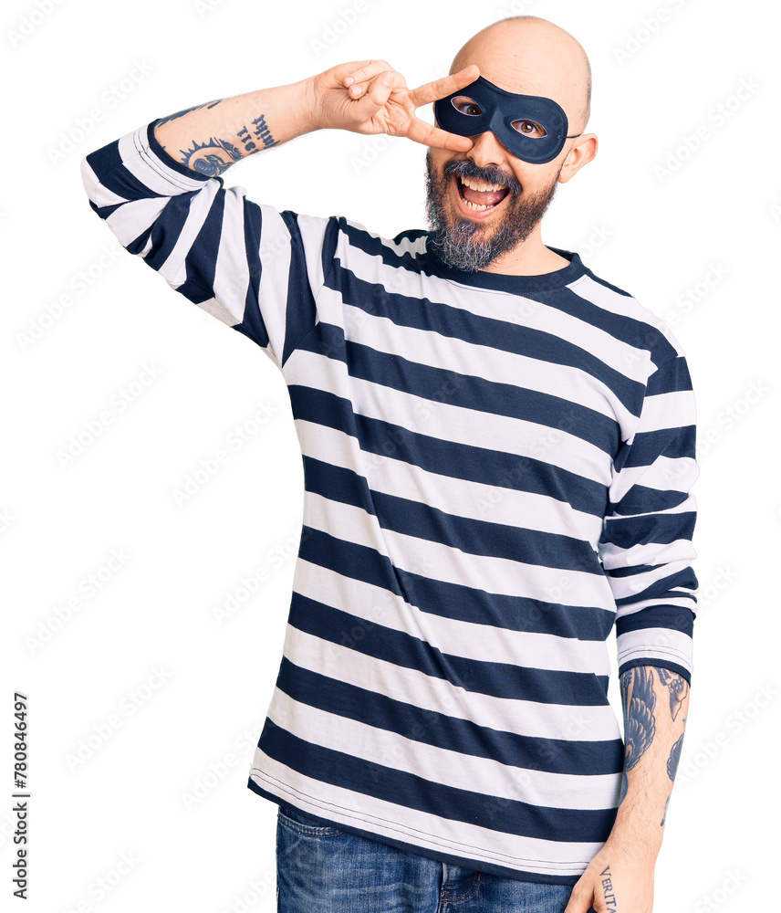 Young handsome man wearing burglar mask doing peace symbol with fingers over face, smiling cheerful showing victory Young handsome man wearing burglar mask doing peace symbol with fingers over face, smiling cheerful showing victory