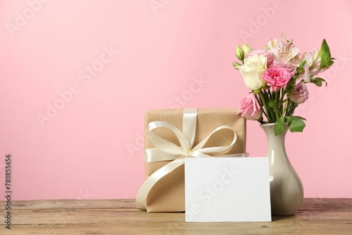 Happy Mother's Day. Gift box, blank card and bouquet of beautiful flowers in vase on wooden table against pink background. Space for text