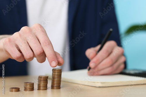 Financial savings. Man stacking coins while writing down notes at wooden table, closeup