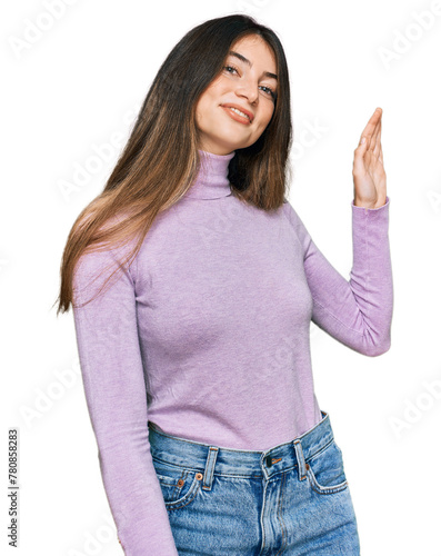 Young beautiful teen girl wearing turtleneck sweater waiving saying hello happy and smiling, friendly welcome gesture