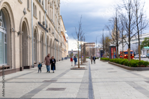 Pedestrian zone of the city street. Defocused photograph of city street. Wide sidewalk with trees and few pedestrians. Spring, cloudy weather photo