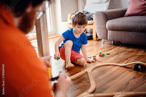 A young boy sits attentively on the floor, engaging with a colorful train set, with an adult's guiding presence nearby, creating a dynamic play environment.