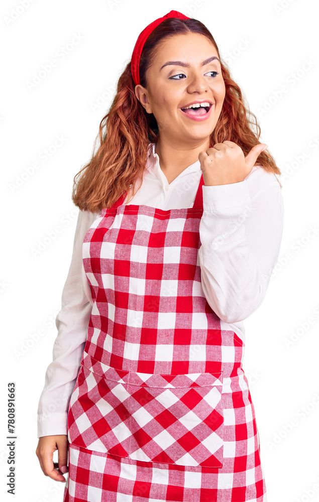 Young latin woman wearing apron smiling with happy face looking and pointing to the side with thumb up. Young latin woman wearing apron smiling with happy face looking and pointing to the side with thumb up.