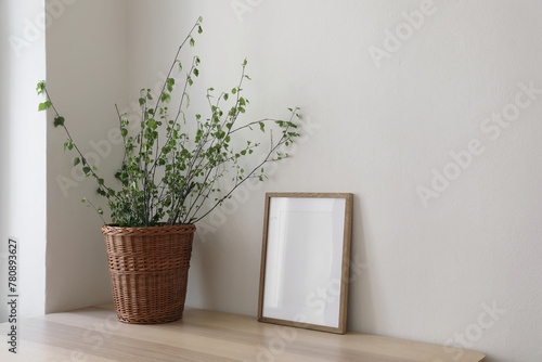 Spring, Easter still life. Elegant Scandinavian living room, home office. Empty vertical wooden picture frame mockup on desk, table. Wicker willow basket with green birch tree branches. Side view. © tabitazn