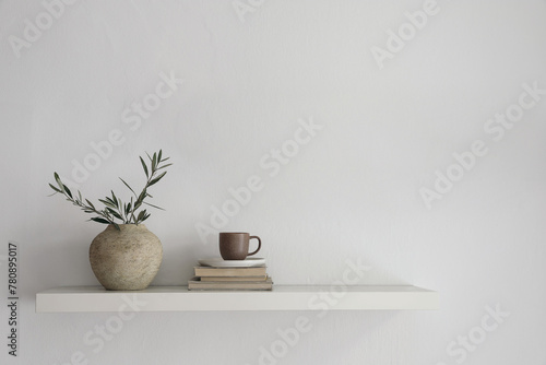 Elegant home still life. Floating shelf. Textured vase with green olive tree branches and old books. Cup of coffee, tea. Modern Mediterranean appartement. White wall background. Interior indoor mockup