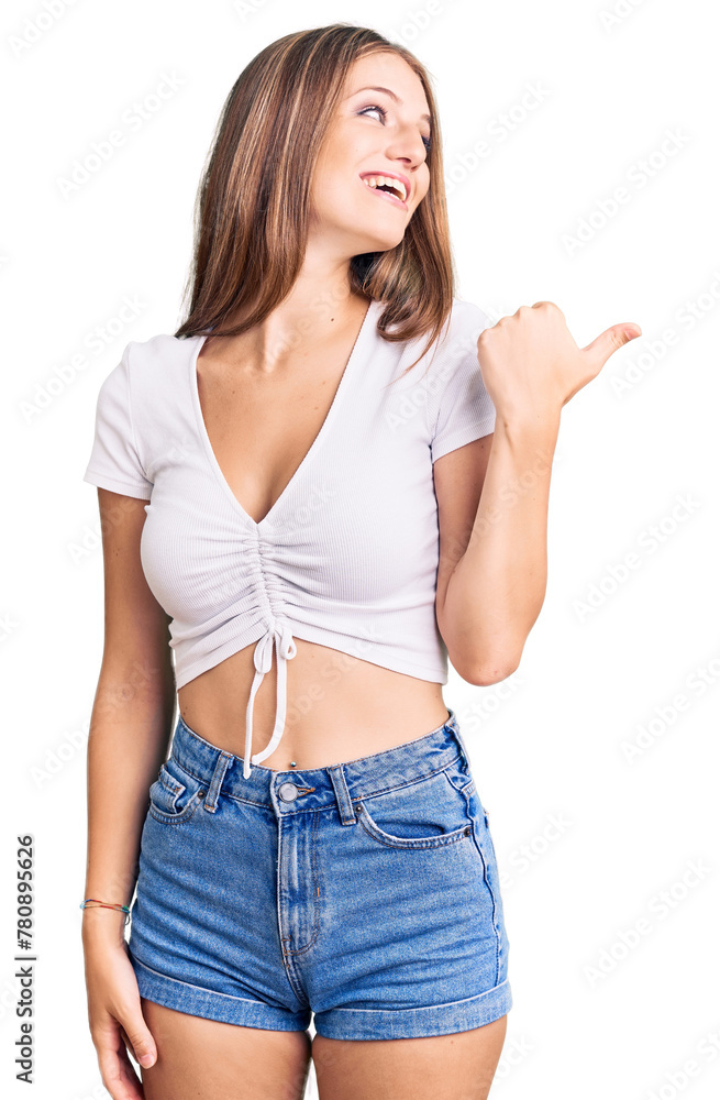 Young beautiful blonde woman wearing casual white tshirt smiling with happy face looking and pointing to the side with thumb up. Young beautiful blonde woman wearing casual white tshirt smiling with happy face looking and pointing to the side with thumb up.