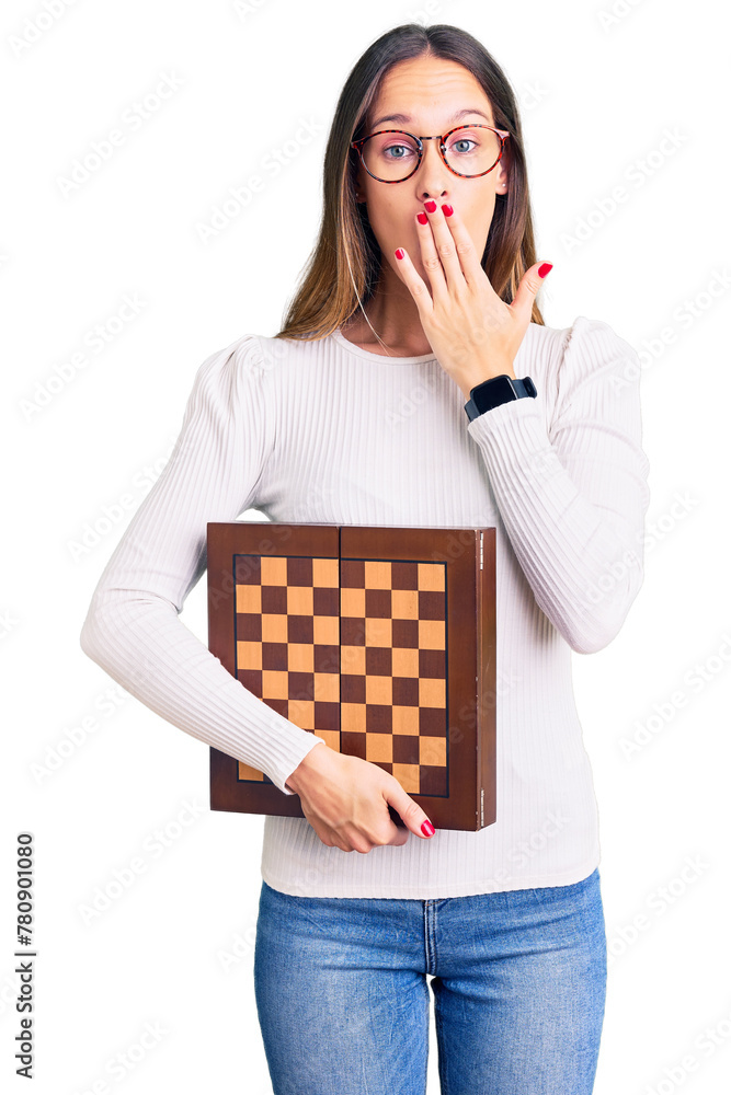 Beautiful brunette young woman holding chess covering mouth with hand, shocked and afraid for mistake. surprised expression
