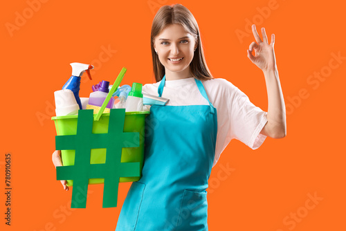 Female janitor holding cleaning supplies, hashtag sign and showing OK gesture on color background