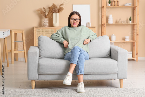 Beautiful young happy Asian woman in stylish eyeglasses sitting on sofa at home © Pixel-Shot