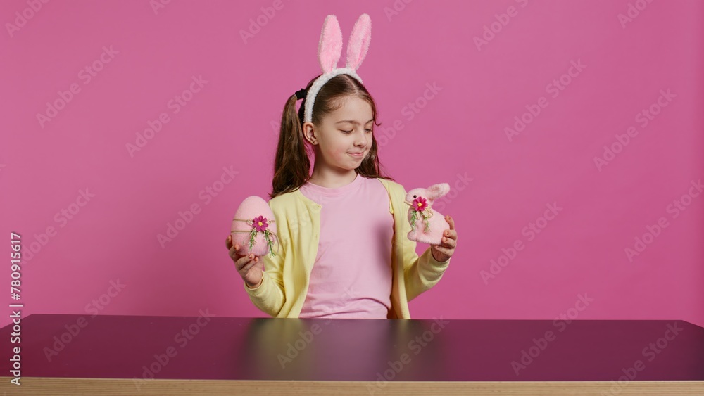 Fototapeta premium Small cheerful girl showing colorful decorated ornaments on camera, creating handcrafted festive arrangements. Young child feeling excited about easter sunday, holds rabbit and egg. Camera B.