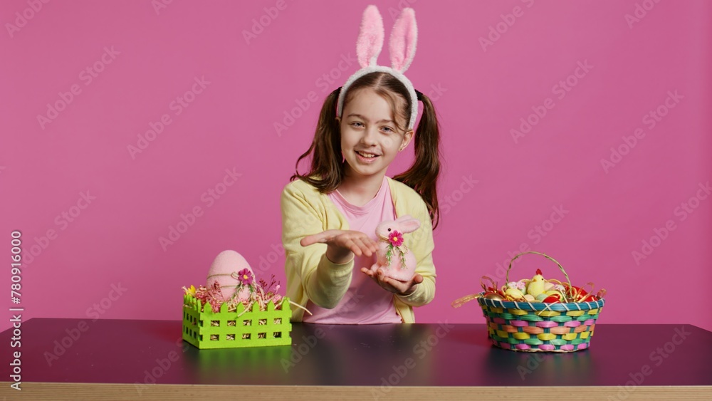 Obraz premium Young schoolgirl blowing air kisses in front of camera while she creates festive lovely arrangements on stuffed rabbit toy. Cheerful young child being excited about easter celebration. Camera B.