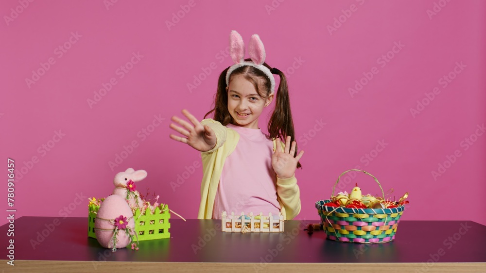 Obraz premium Joyful schoolgirl with bunny ears waving hello in front of camera, sitting at a table with festive decorations and arrangements for easter sunday celebration. Young excited child. Camera B.