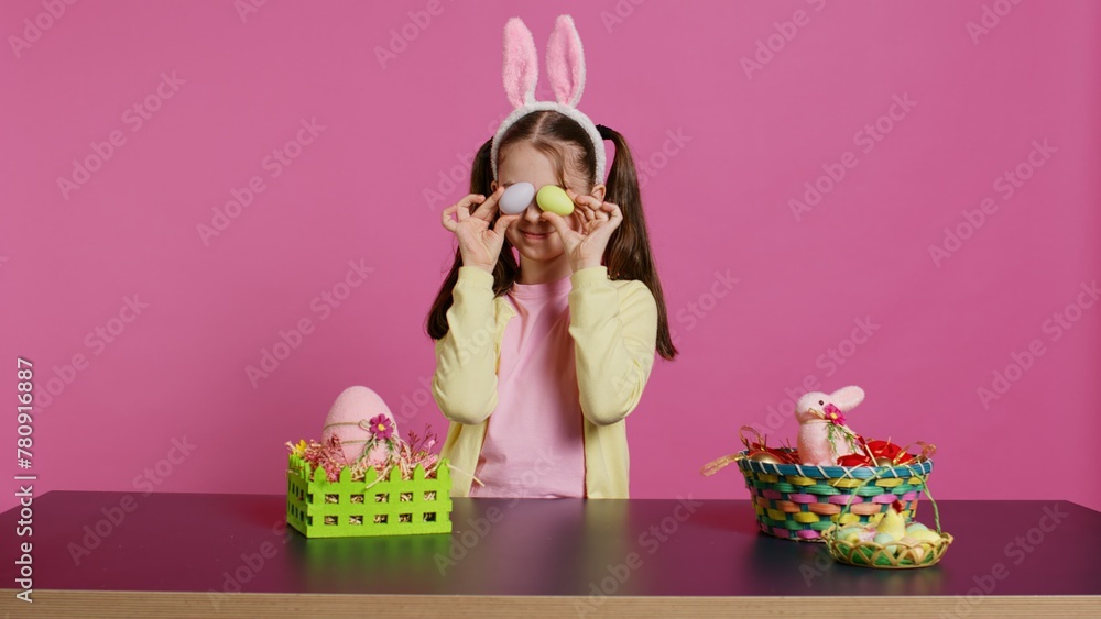 Obraz premium Enthusiastic young girl playing peek a boo in front of camera, using painted easter eggs against pink background. Joyful lovely toddler feeling excited about spring holiday festivity. Camera B.