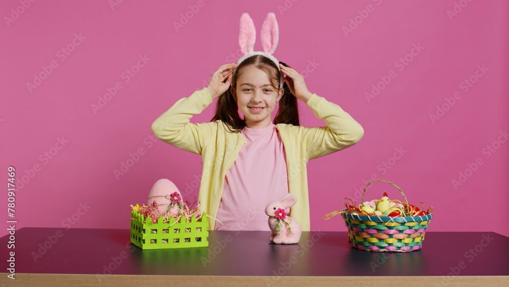 Fototapeta premium Smiling confident toddler placing bunny ears on her head and waving, saying hello against pink background. Cute joyful child creating decorations for easter sunday holiday event. Camera B.