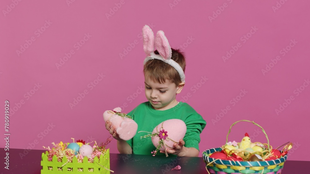 Obraz premium Small pleased child presenting hid handcrafted ornaments created in preparation of easter holiday celebration. Young boy with bunny ears showing painted decorations, a rabbit and an egg. Camera A.