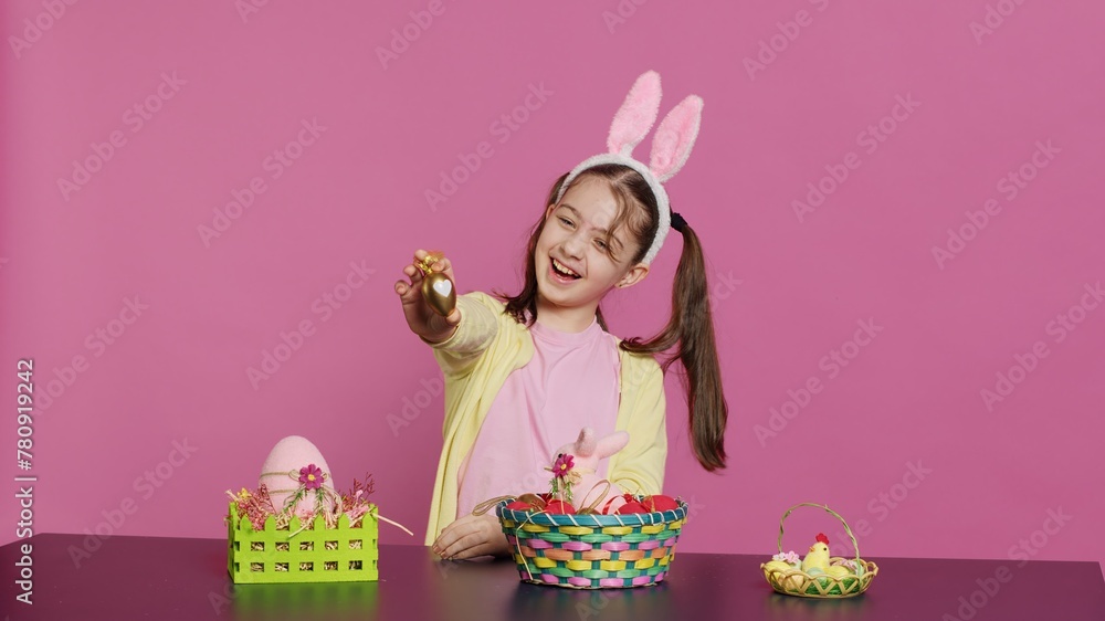 Fototapeta premium Excited young girl arranging painted eggs in a basket to prepare for easter holiday celebration, creating festive arrangements. Playful happy toddler with bunny ears, creative activity. Camera A.