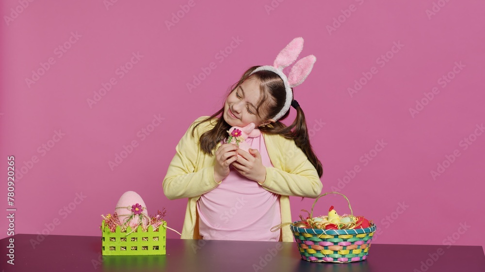 Naklejka premium Cheerful smiling girl hugging her stuffed rabbit toy and egg, feeling proud about her handcrafted painted easter decorations. Young toddler with bunny ears and pigtails, festive ornaments. Camera A.
