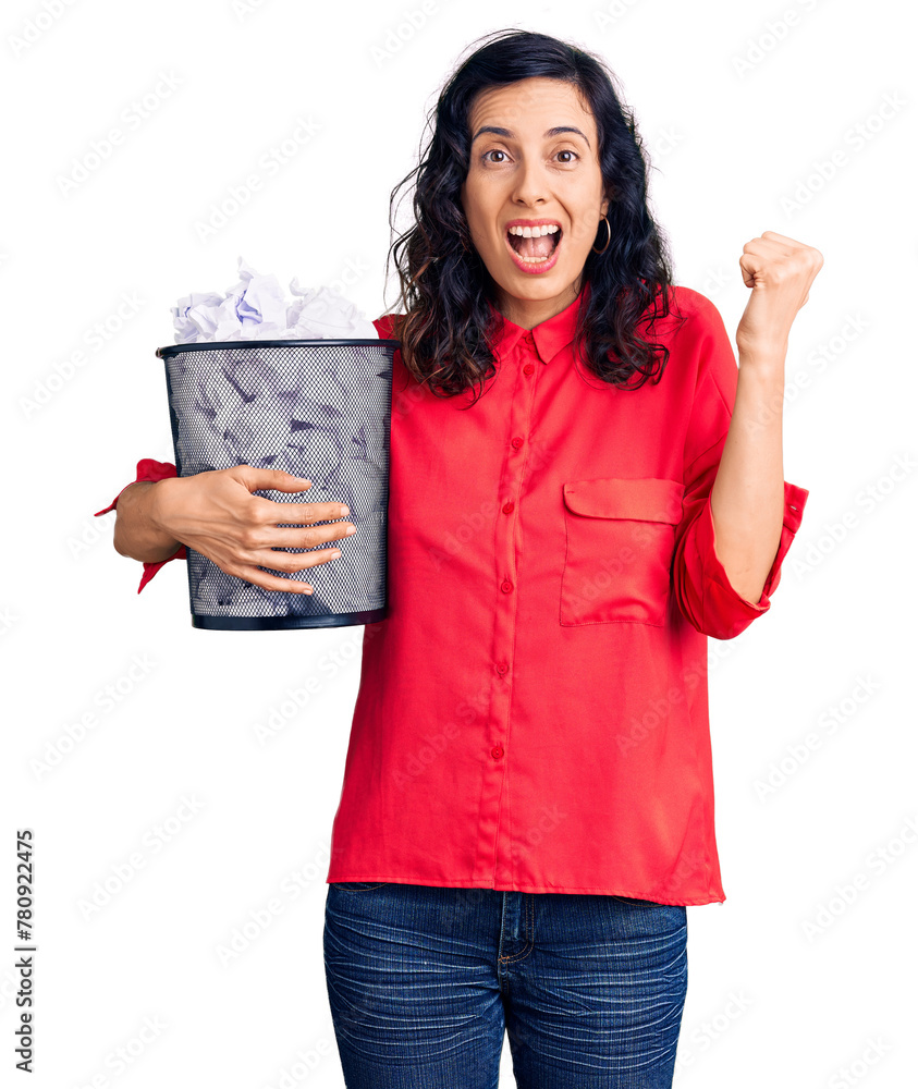 Obraz premium Young beautiful hispanic woman holding paper bin full of crumpled papers screaming proud, celebrating victory and success very excited with raised arms