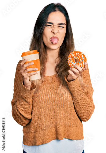 Beautiful hispanic woman eating doughnut and drinking coffee sticking tongue out happy with funny expression.