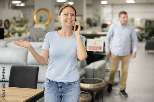 Cheerful young woman talking emotionally on mobile phone while strolling through furnishing showroom and examining pieces of furniture with interest