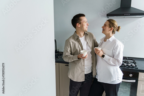 A couple shares a warm, convivial moment in their sleek kitchen, both clad in neutral tones, with mugs in hand, exuding a casual, comfortable domesticity.