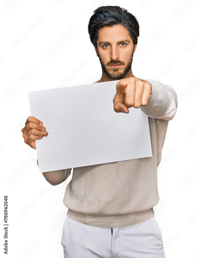 Young hispanic man holding blank empty banner pointing with finger to the camera and to you, confident gesture looking serious Young hispanic man holding blank empty banner pointing with finger to the camera and to you, confident gesture looking serious