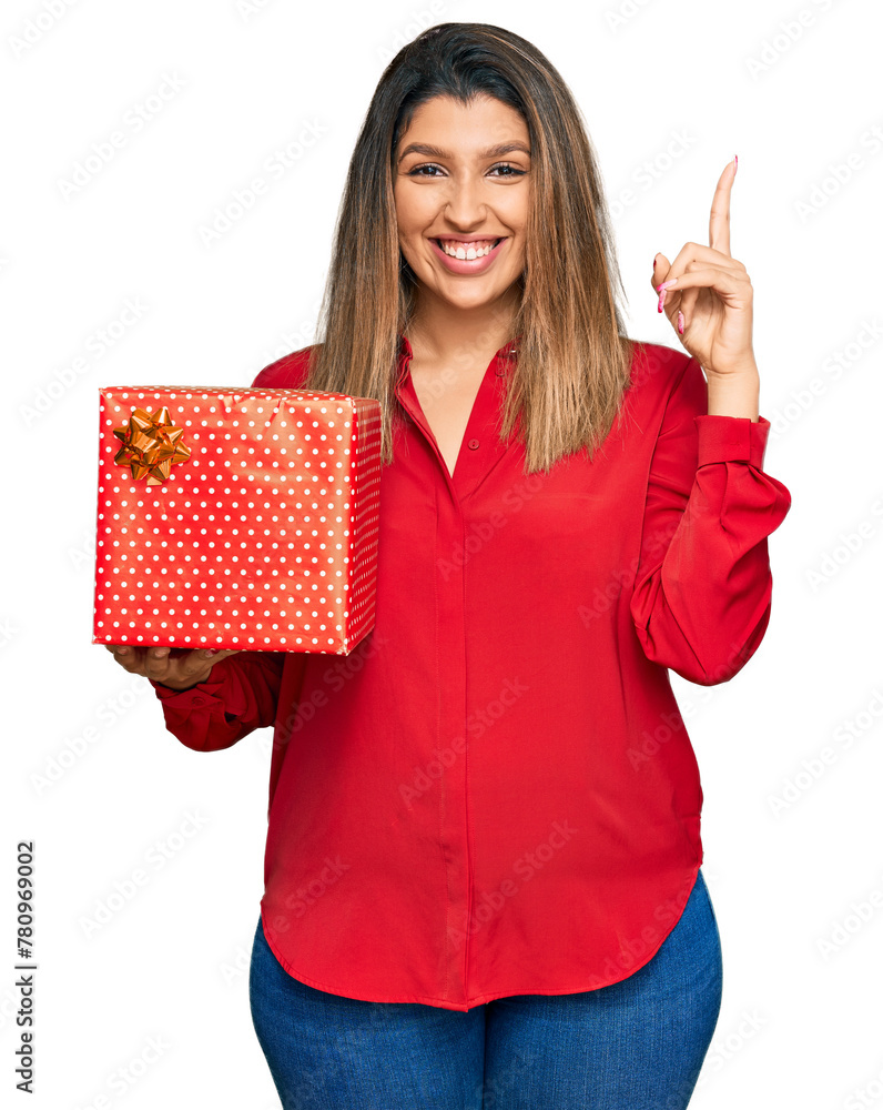 Beautiful brunette woman holding gift surprised with an idea or question pointing finger with happy face, number one Beautiful brunette woman holding gift surprised with an idea or question pointing finger with happy face, number one