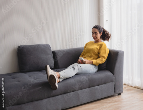 Portrait latin american woman one person smile happy sitting sofa and looking and hand holding use remote control adjust air conditioning inside. on most wonderful vacation day in living room