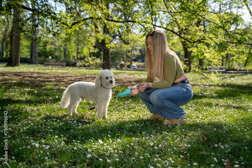 Beautiful girl giving water to her pet poodle in the park 