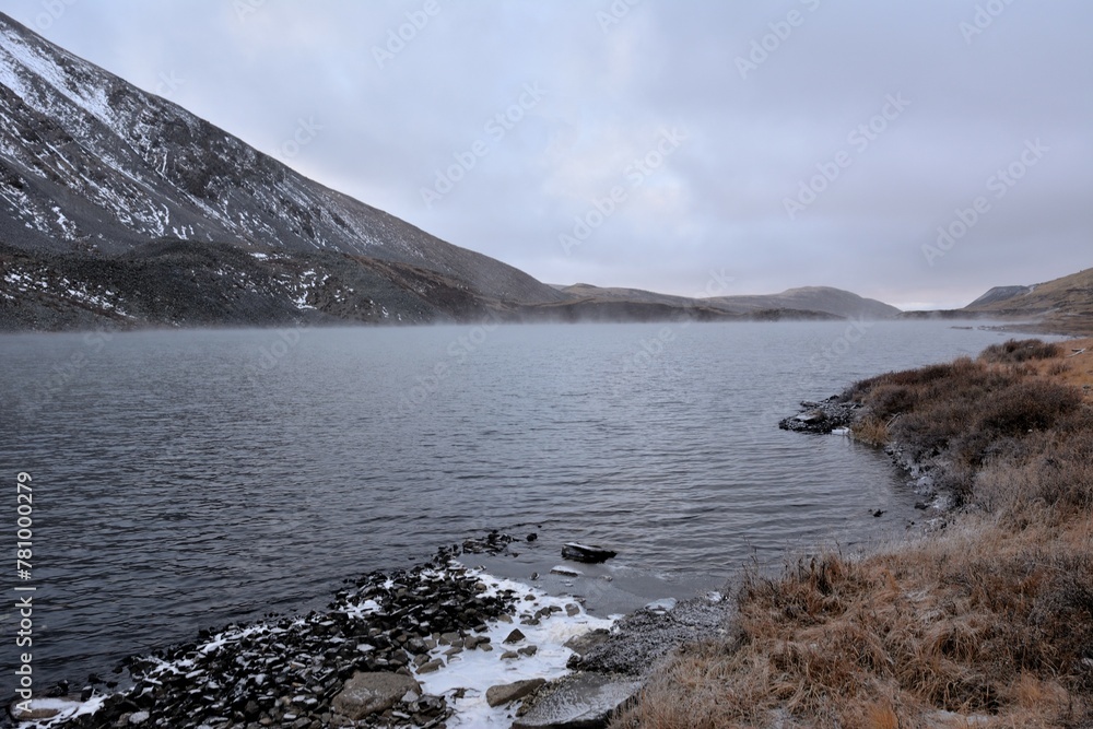 The shore of a large lake, taken by the first ice at the foot of a high snow-covered mountain, on a cloudy autumn morning. The shore of a large lake, taken by the first ice at the foot of a high snow-covered mountain, on a cloudy autumn morning.