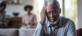 An older African man is sitting on a couch in the living room, looking hurt and lonely. He touches his head, seemingly suffering from a severe headache or recalling painful memories.