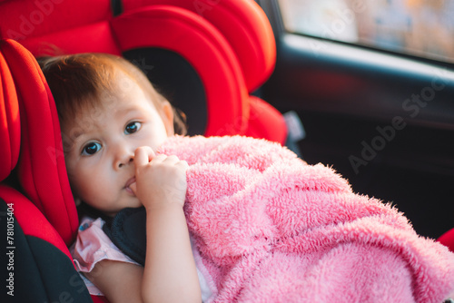 Asian Toddler Girl seats peacefully in her red carseat. She is holding her pink blanket while sucking on her thumb during sunrise or sunset. While looking at the camera.