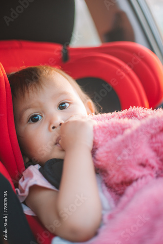 Asian Toddler Girl seats peacefully in her red carseat. She is holding her pink blanket while sucking on her thumb during sunrise or sunset. While looking at the camera.