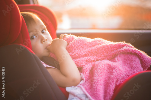 Asian Toddler Girl seats peacefully in her red carseat. She is holding her pink blanket while sucking on her thumb during sunrise or sunset. While looking at the camera.