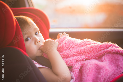 Asian Toddler Girl seats peacefully in her red carseat. She is holding her pink blanket while sucking on her thumb during sunrise or sunset. While looking at the camera.