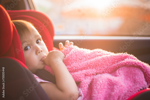 Asian Toddler Girl seats peacefully in her red carseat. She is holding her pink blanket while sucking on her thumb during sunrise or sunset. While looking at the camera.