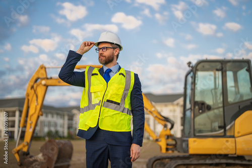 Construction builder investor. Man investor in front of construction site. Successful handsome man standing at modern home building construction. Portrait of midlle aged investor in suit and helmet. photo