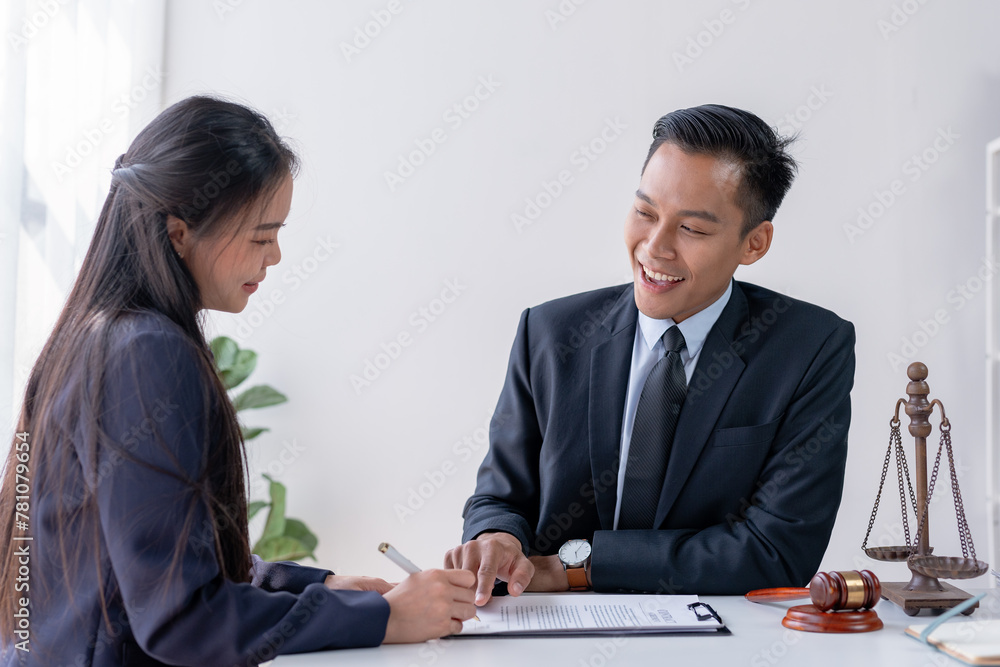 Lawyer smiles encouragingly as a client signs documents, scales of justice and gavel in the foreground. Lawyer smiles encouragingly as a client signs documents, scales of justice and gavel in the foreground.
