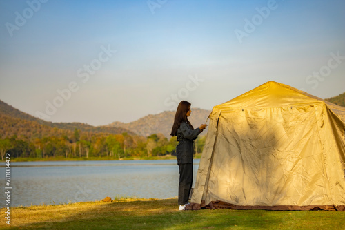 Asian woman traveler setting up luxury tent during sunset camping in forest near lake. Landscape with sunlight shining through orange clouds. Activity and lifestyle on holiday, eco-friendly adventure.