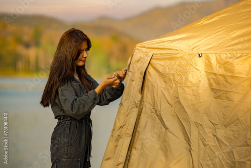 Asian woman traveller installation set up of a tarp or tent made of rip-stop fabric at the mountains near of the lake in the nature on vacation holiday with beautiful sunset background.