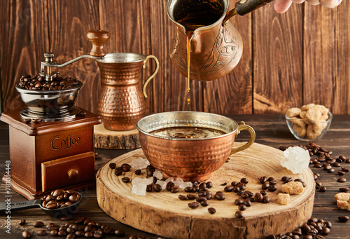 Pouring coffee from coffee maker into copper cup, an antique coffee grinder and copper milk jug on wooden background