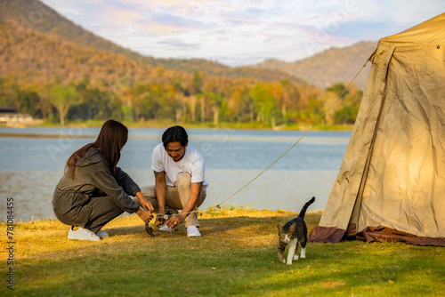 Asian couple camping for the weekend in the woods near the river, Camping Holiday In Countryside, Young couple setting up tent outdoors together while cat walking at green grass lawn near tent.