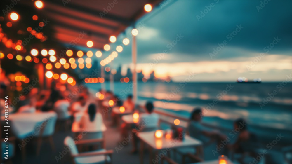 Obraz premium Blurred image shows bokeh of lights of a restaurant on a promenade by the sea. Unknown people are sitting at tables by candlelight. The sea, sunset and a ship can be seen in the background.